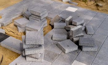 Gray paving stones being laid on a sandy surface, some stacked nearby.