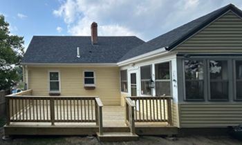 Back of a yellow house with a wooden deck and sunroom. Blue sky, a brick chimney, and green trees are visible.