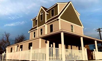 Tan and green Victorian house with a porch and white picket fence, set against a cloudy sky.