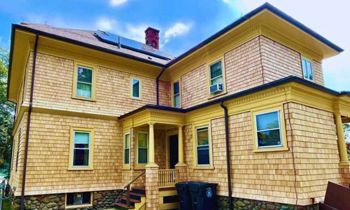 Yellow-trimmed house with cedar shake siding; solar panels on roof, blue sky background.