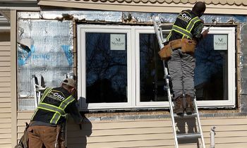 Two workers installing a window on a house. One on a ladder, applying sealant. Both wear reflective vests.