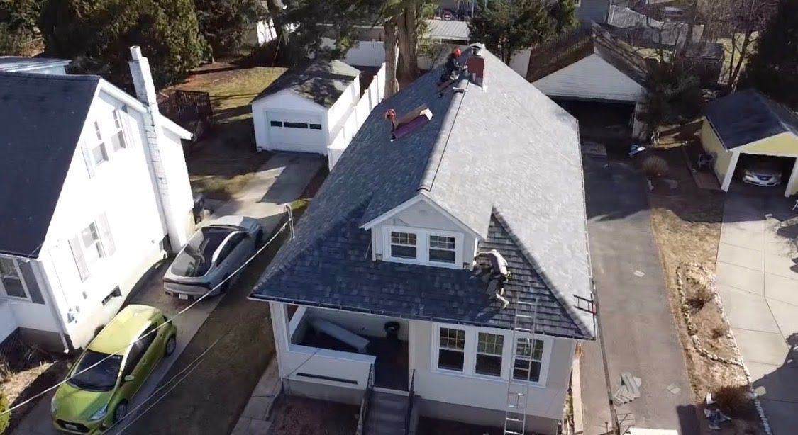 Aerial view of a white house with gray roof and driveway, surrounded by trees and neighboring homes.