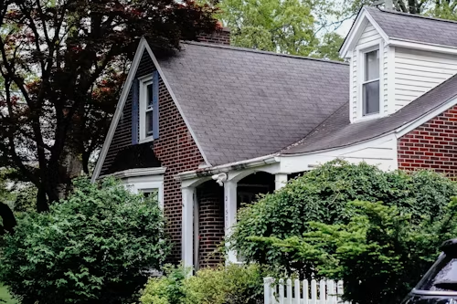 Brick house with gray roof, white porch, and dense green shrubs along the front yard