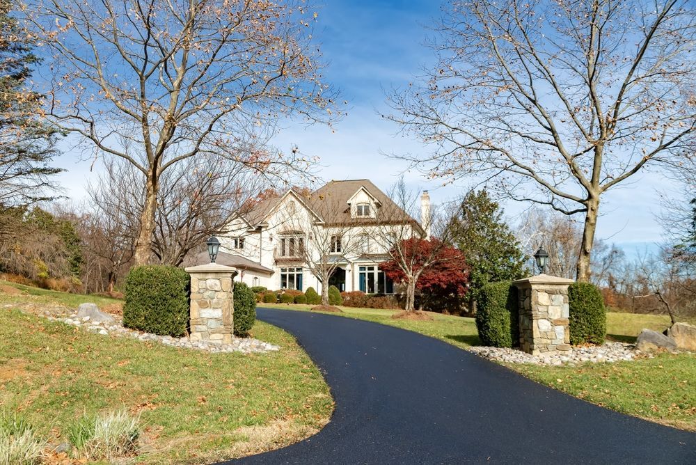 A large, stone house with a curved driveway, flanked by trees and pillars.