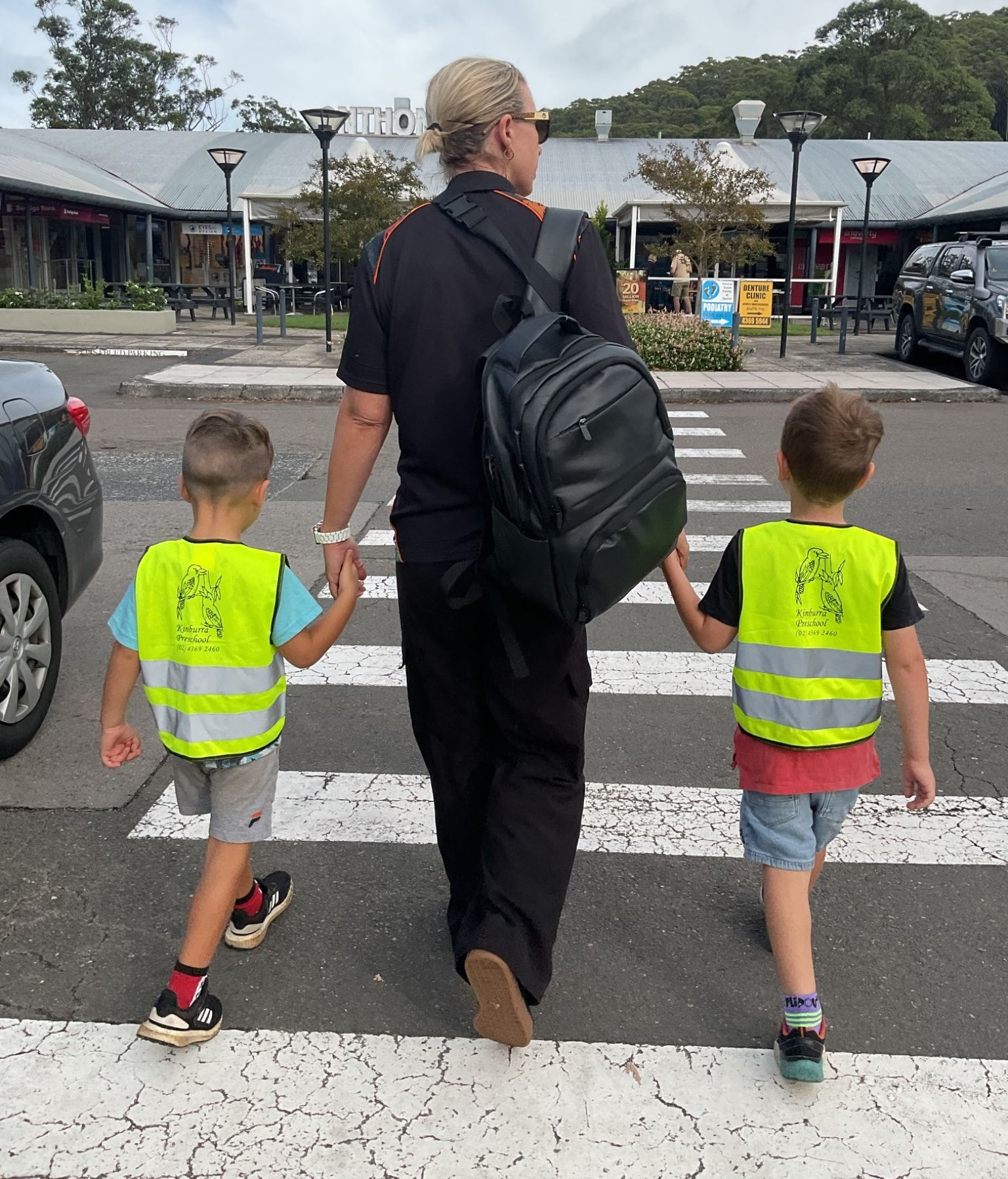 A person wearing a backpack and holding hands with two children crossing a crosswalk. — Kinburra Preschool (Kincumber) in Kincumber, NSW