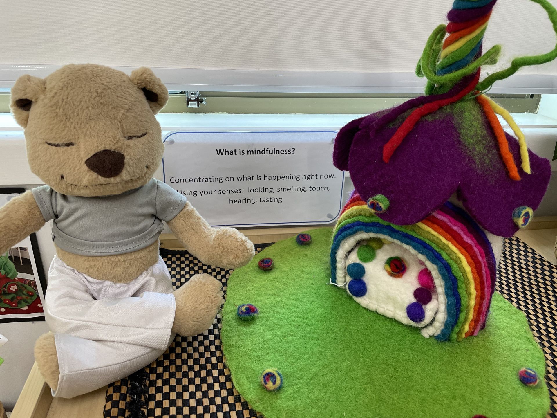 Teddy bear meditating next to a felt rainbow house with a rainbow arch and colourful swirls — Kinburra Preschool (Kincumber) in Kincumber, NSW