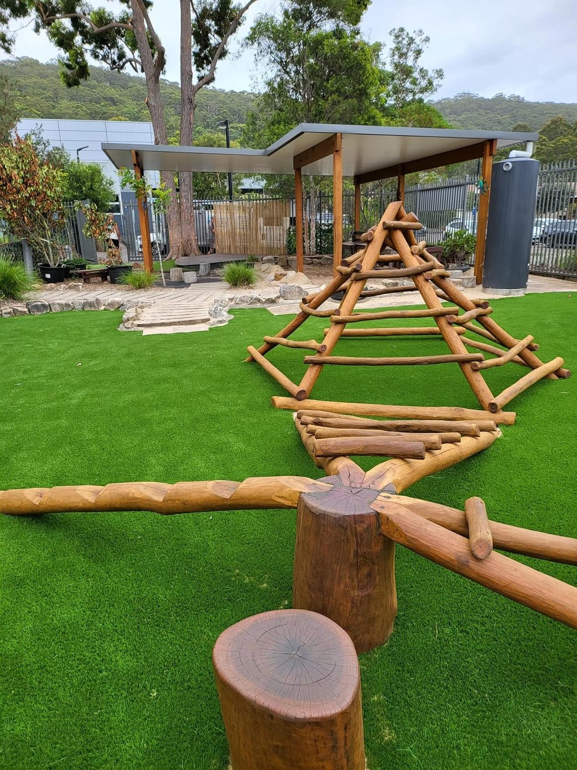 Playground with wooden structures on green turf, shed in background. — Kinburra Preschool (Kincumber) in Kincumber, NSW