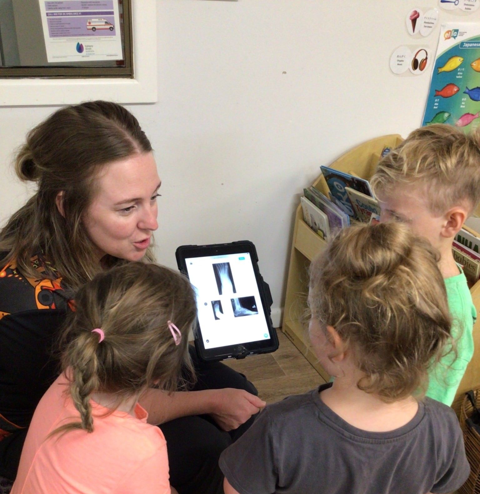 Woman shows x-ray on tablet to three children, all looking intently. Indoors. — Kinburra Preschool (Kincumber) in Kincumber, NSW