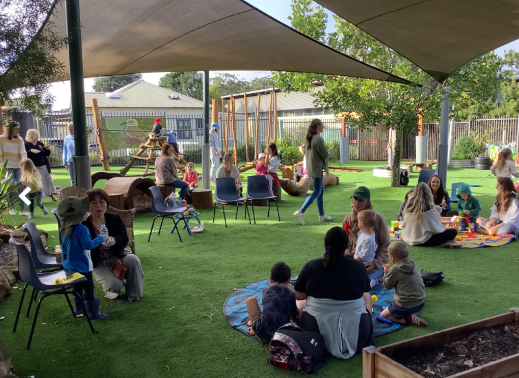 People and children gathered outdoors on artificial turf under a sunshade. — Kinburra Preschool (Kincumber) in Kincumber, NSW