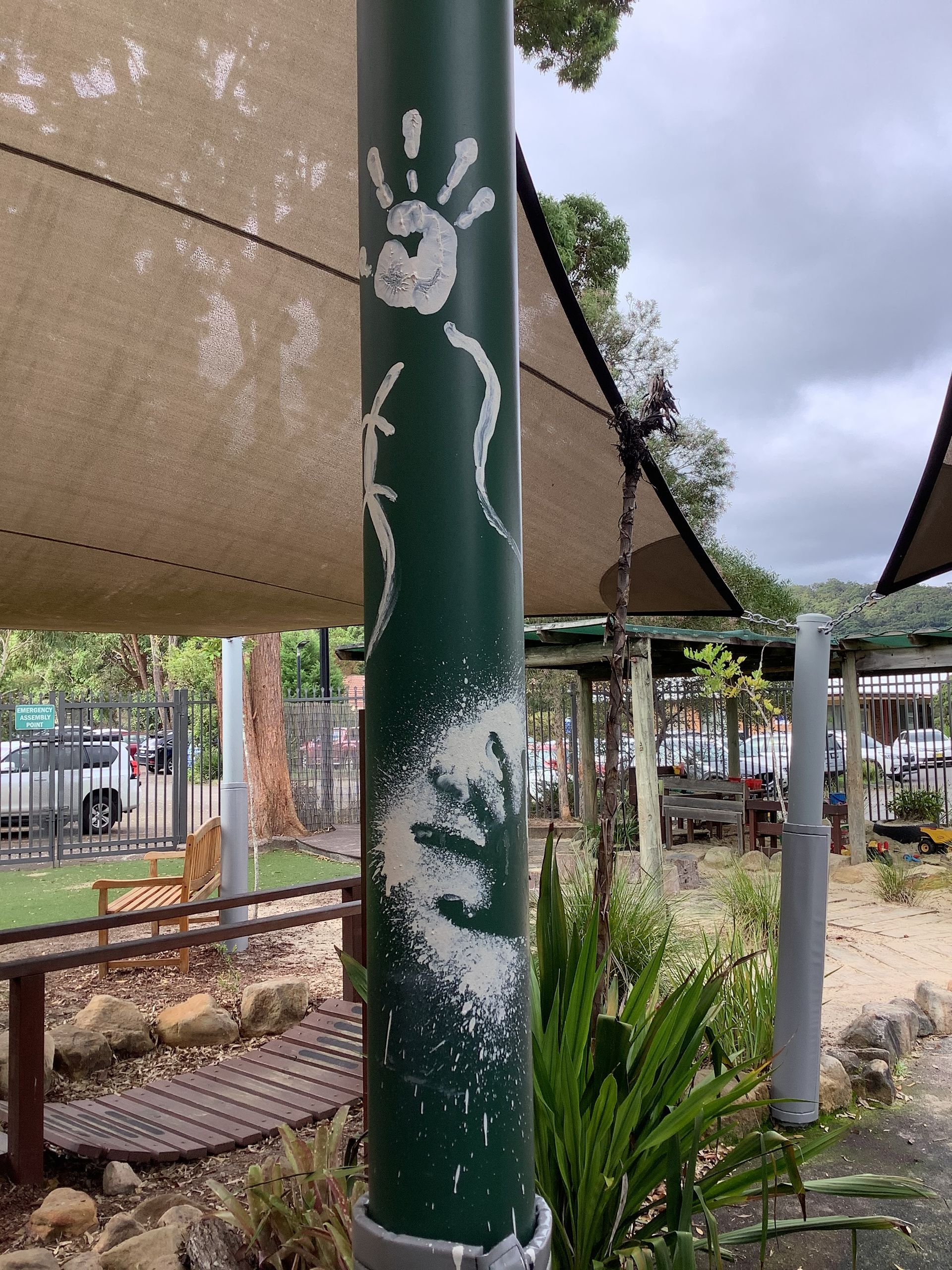Green pole with white handprints and abstract design; outdoor setting with shade sails. — Kinburra Preschool (Kincumber) in Kincumber, NSW