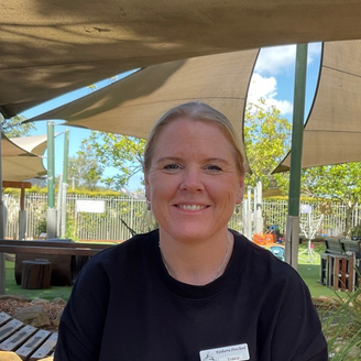 Smiling person seated outdoors under a shade canopy, wearing a black shirt.