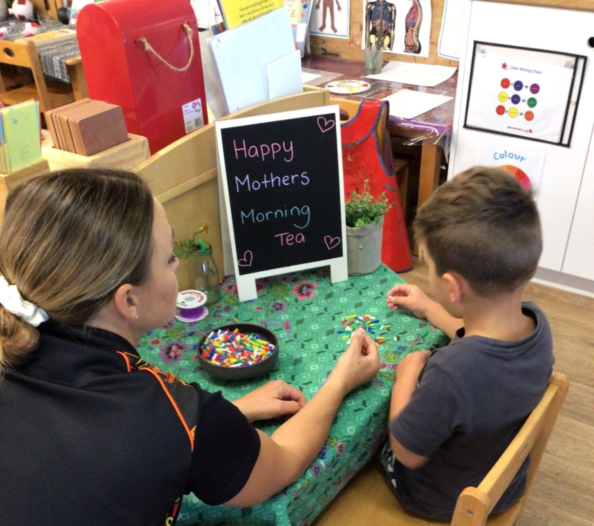 Woman and boy crafting at table, Happy Mother's Day sign visible. Indoors with colorful beads. — Kinburra Preschool (Kincumber) in Kincumber, NSW