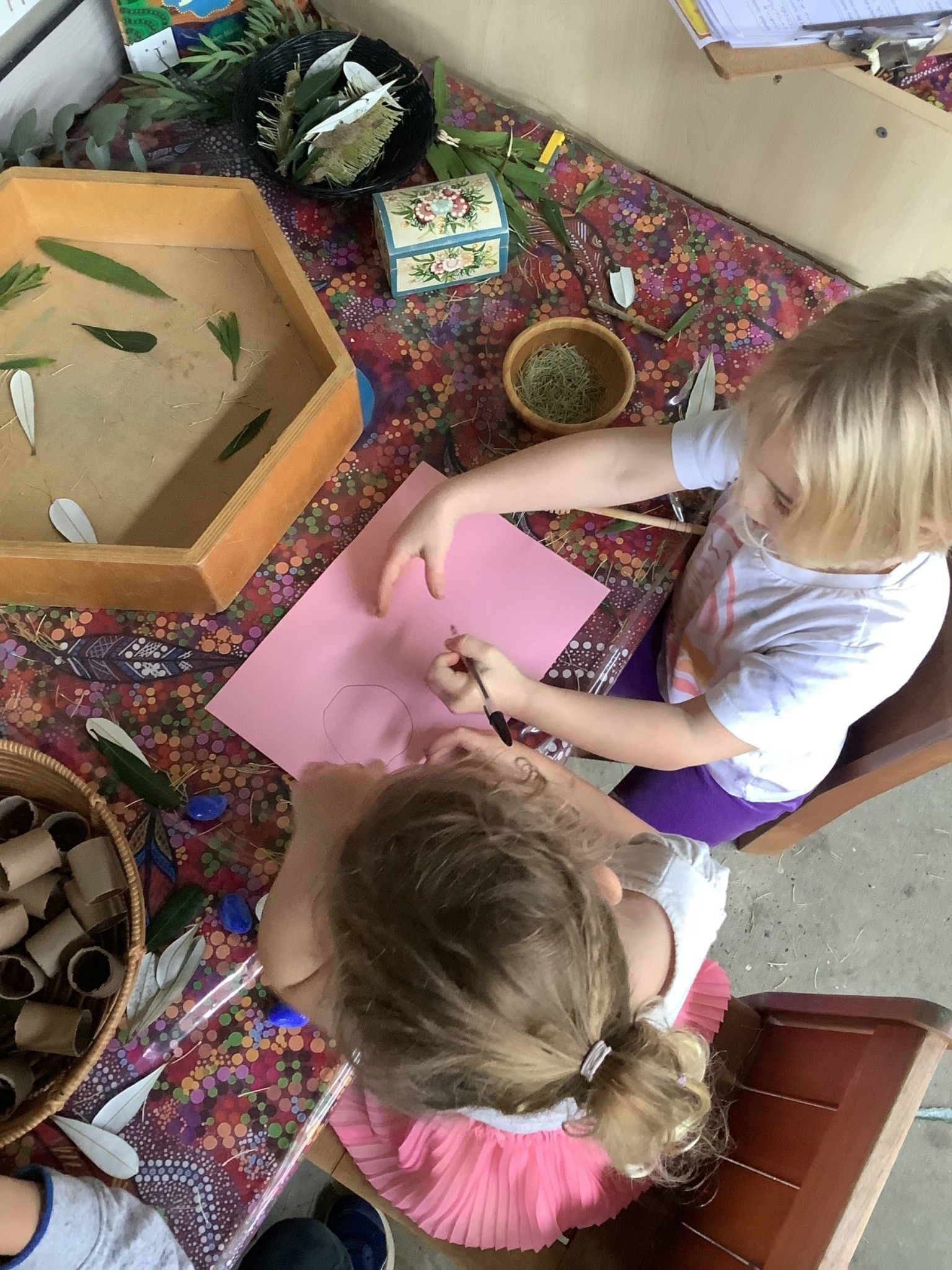 Two young children drawing on pink paper at a table. Various natural items and art supplies surround them. — Kinburra Preschool (Kincumber) in Kincumber, NSW