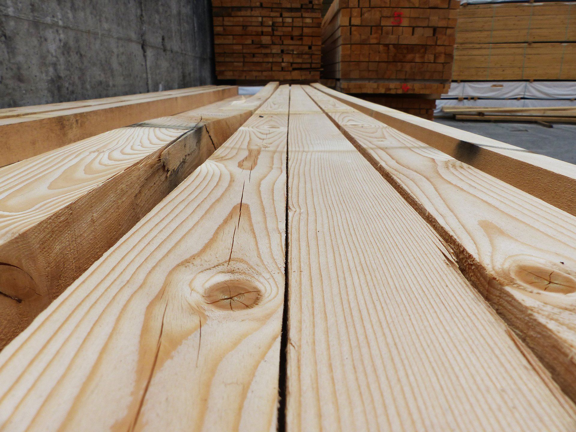 Primo piano di assi di legno impilate in un cantiere, che mostrano le venature chiare del pino e un muro di mattoni sullo sfondo.