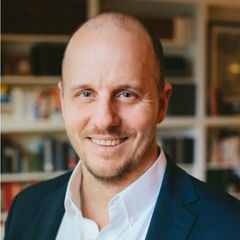 Smiling person in a white shirt and dark blazer in front of a blurred bookshelf background