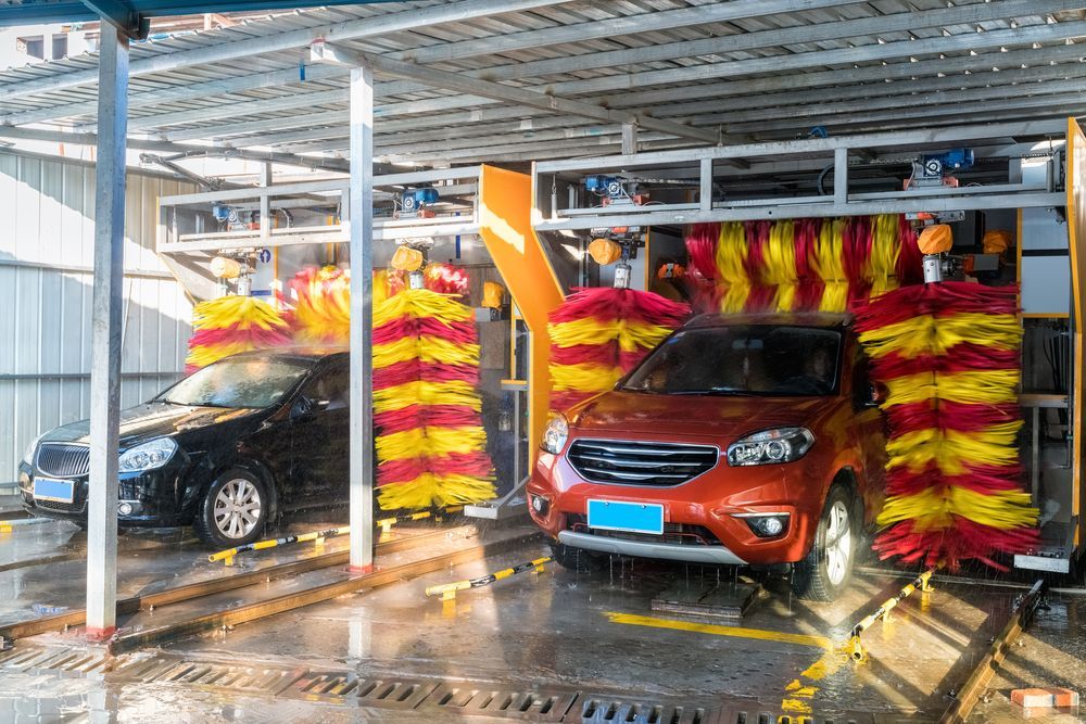 Two cars being cleaned by automated red and yellow brushes in an open-air drive-through car wash station.
