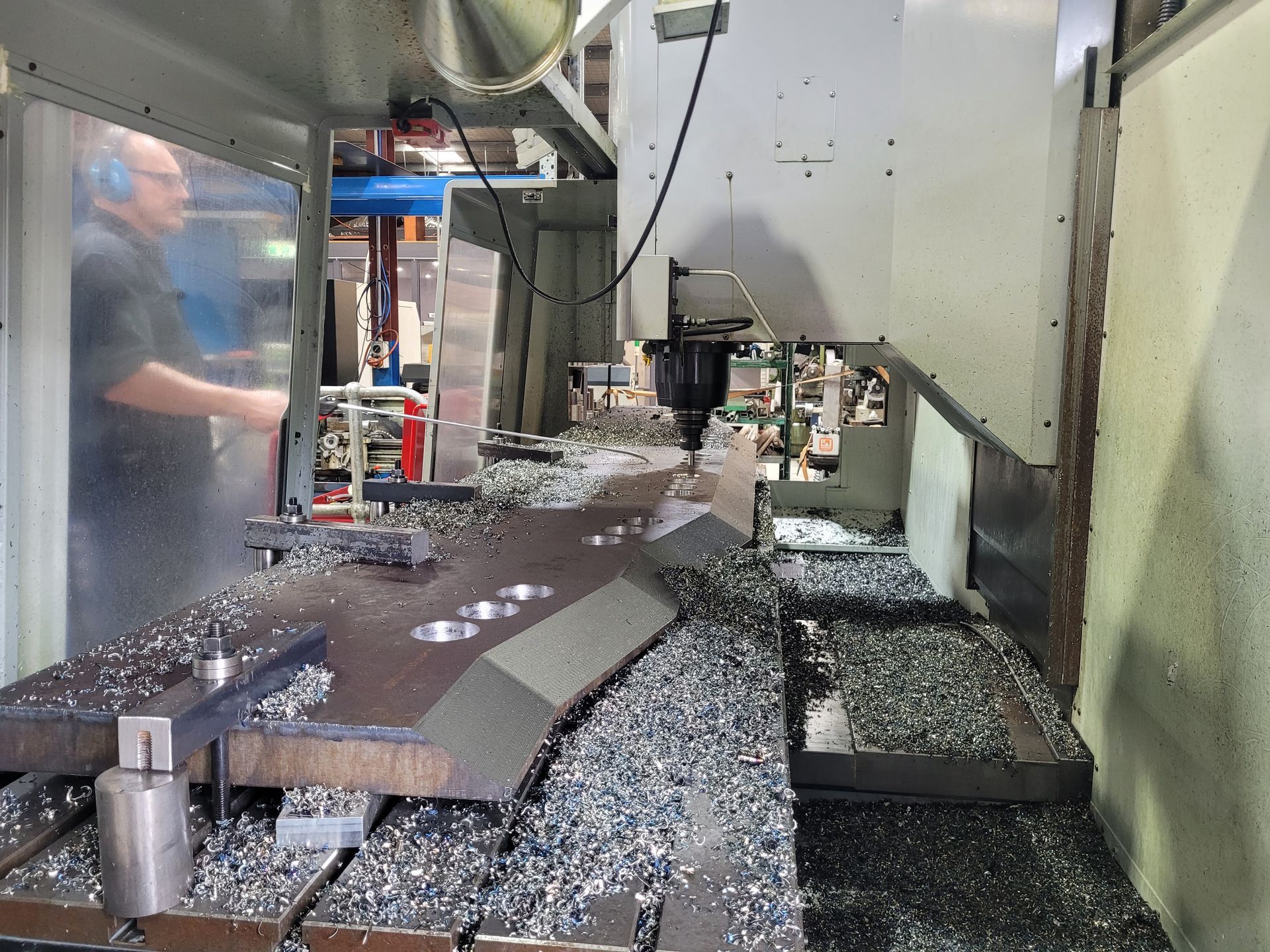 CNC milling machine cutting metal chips, with a worker visible behind the safety panel. — Real Engineering In Gosford, NSW