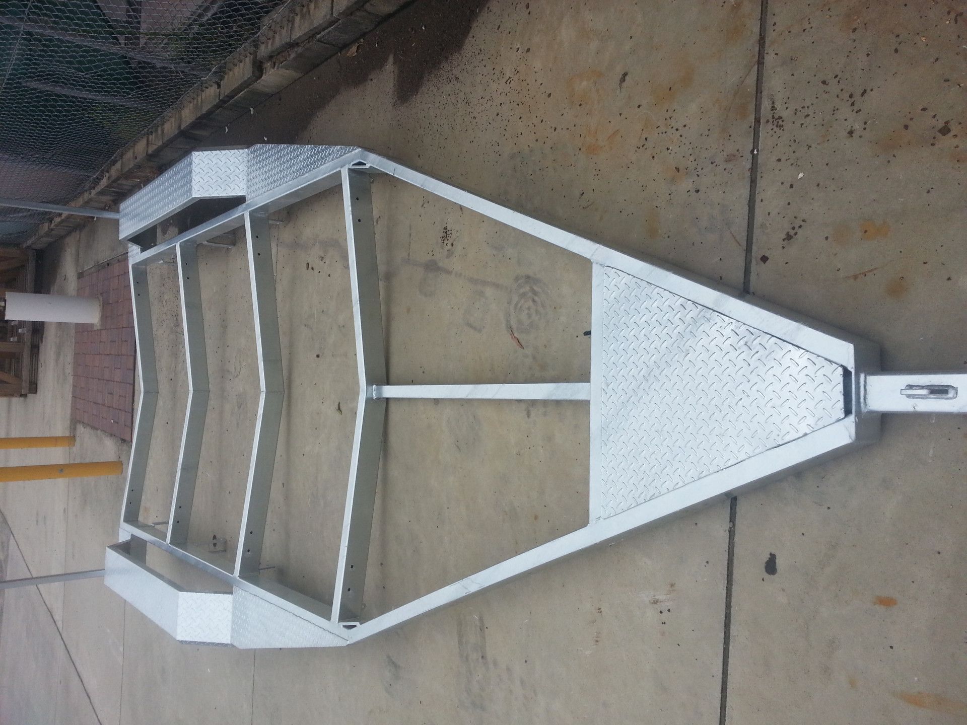 A galvanized steel trailer frame with diamond-plate metal sections, viewed from above on a concrete surface.