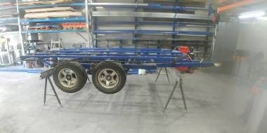 A blue metal dual-axle trailer frame sits on two black sawhorses inside a large warehouse or workshop.