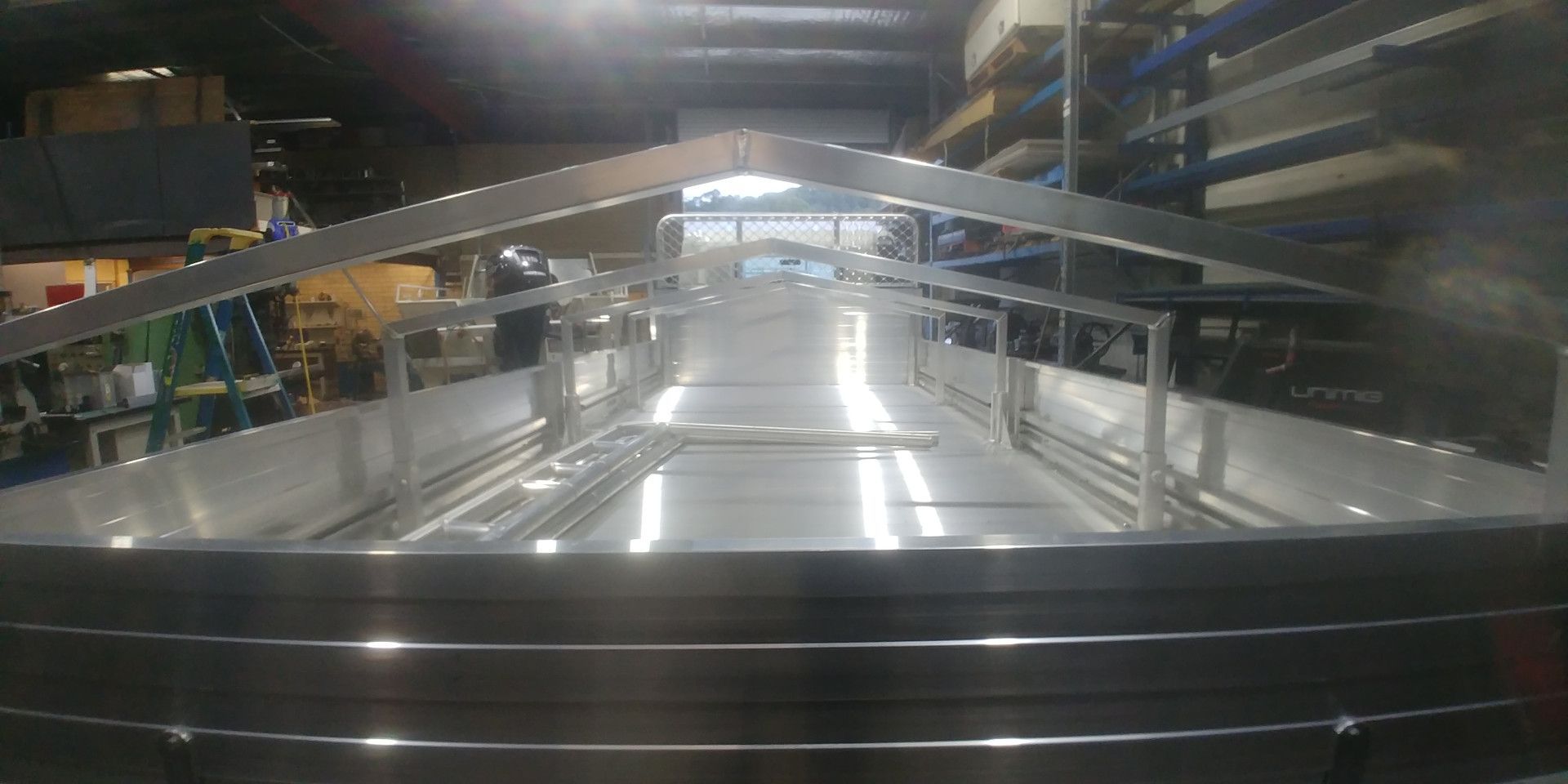 A view from the back of an empty, silver aluminum truck bed, framed by a metal support structure inside a workshop.