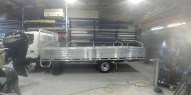A white flatbed truck parked inside a warehouse or workshop, with a large boat motor positioned in the foreground.