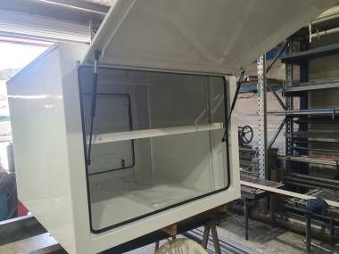 A white, rectangular metal storage box sits in a workshop, its hinged door open and supported by two gas struts.