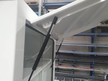 A close-up view of two black gas struts holding open the white lid of a metal storage cabinet in a warehouse.