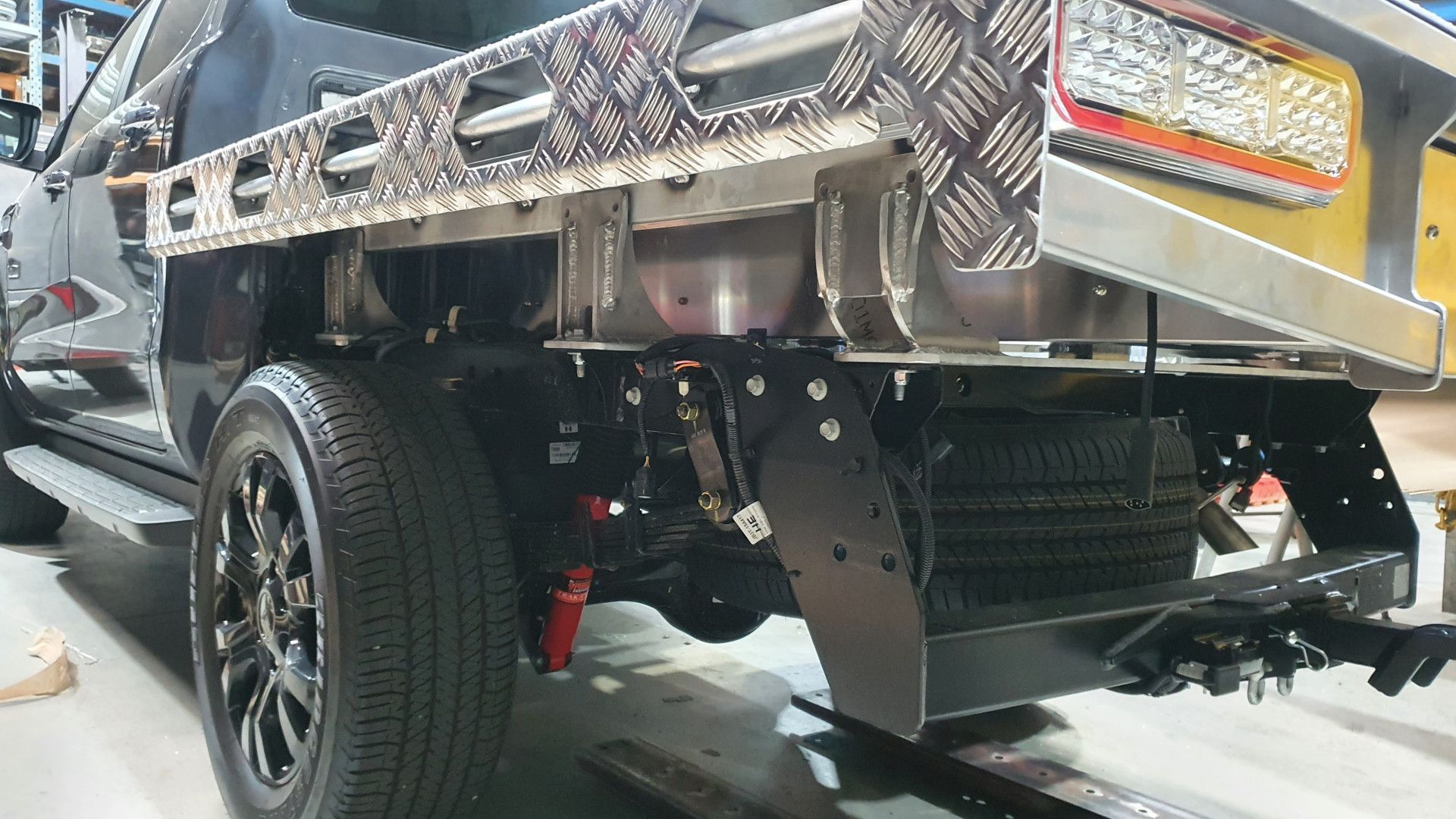 The rear end of a black pickup truck with a metal tray, showing the suspension, spare tire, and towing hitch.