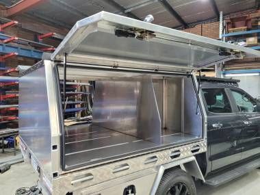 A silver aluminum truck canopy with an open side door, showcasing interior storage partitions, parked in a workshop.