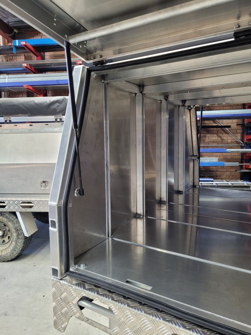 An open aluminum truck canopy featuring internal support pillars, a flat base, and a gas strut for the door hinge.