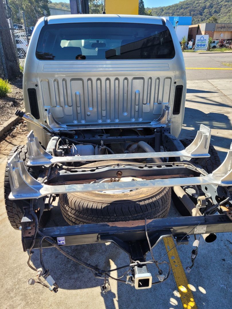 A silver pickup truck chassis with the cargo bed removed, exposing the spare tire, metal frame, and hitch on a sunny day.