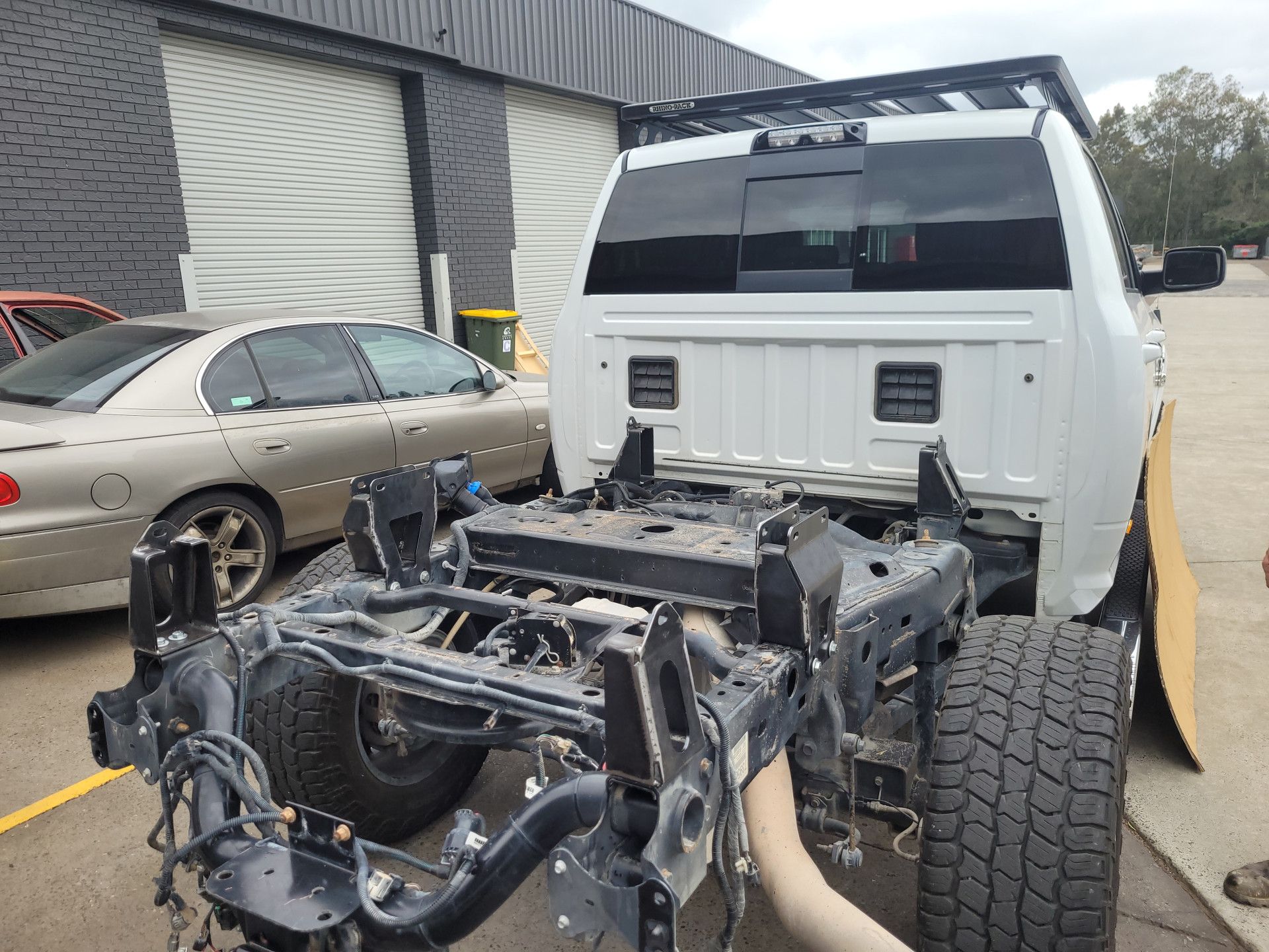 A white truck chassis missing its rear cargo bed, parked outside a building next to a tan sedan.