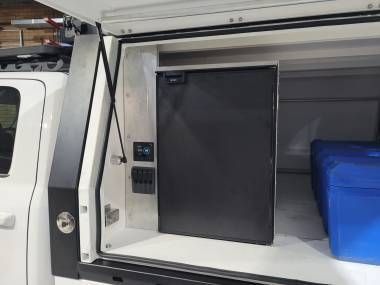 A black refrigerator installed in the open cargo compartment of a white truck, with a control panel and switches nearby.