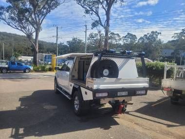 A white utility truck with a custom service canopy, roof rack, and spare tire mounted on the rear, parked in a lot.