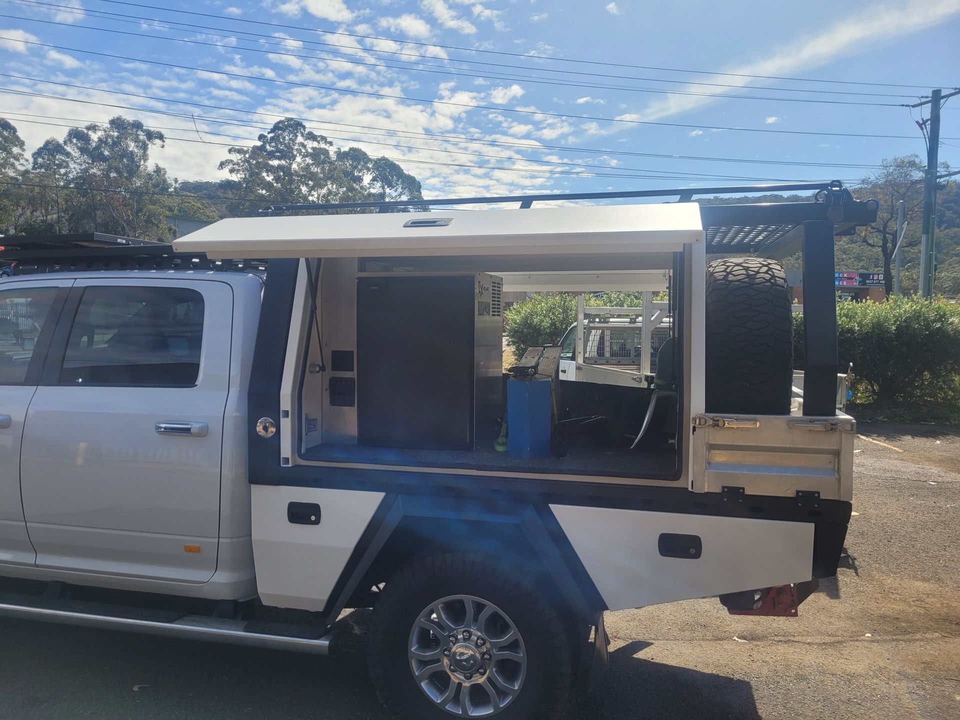 Side view of a white utility truck with a custom open canopy containing a refrigerator and spare tire outdoors.