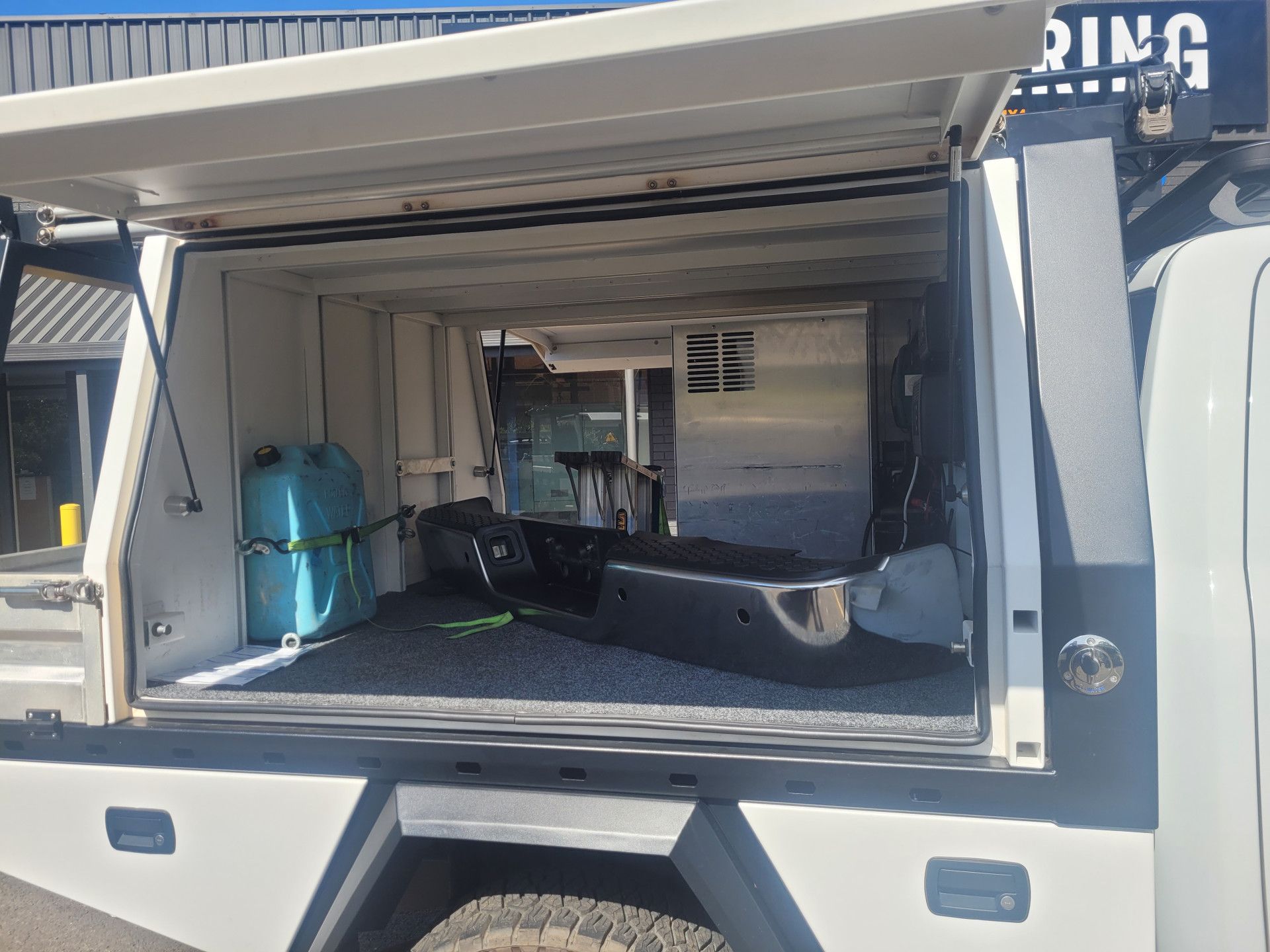 The side compartment of a white truck canopy is open, revealing a blue water container and a black bumper stored inside.