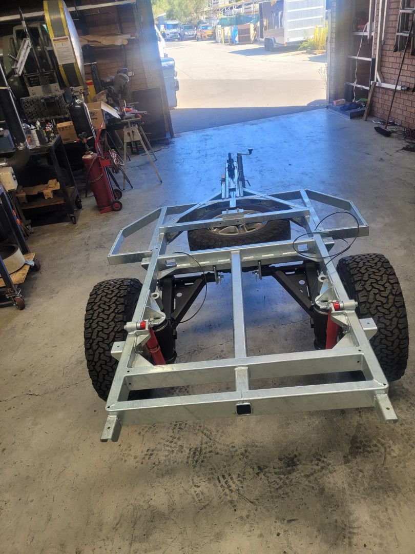 A galvanized metal trailer frame with off-road tires and suspension components resting on a workshop floor.