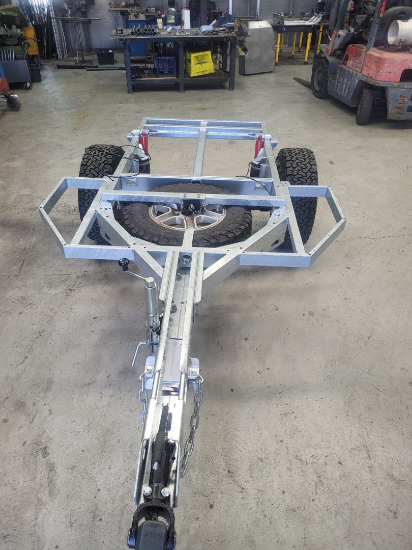 A galvanized metal trailer frame with two knobby tires and a spare wheel, positioned in a workshop.