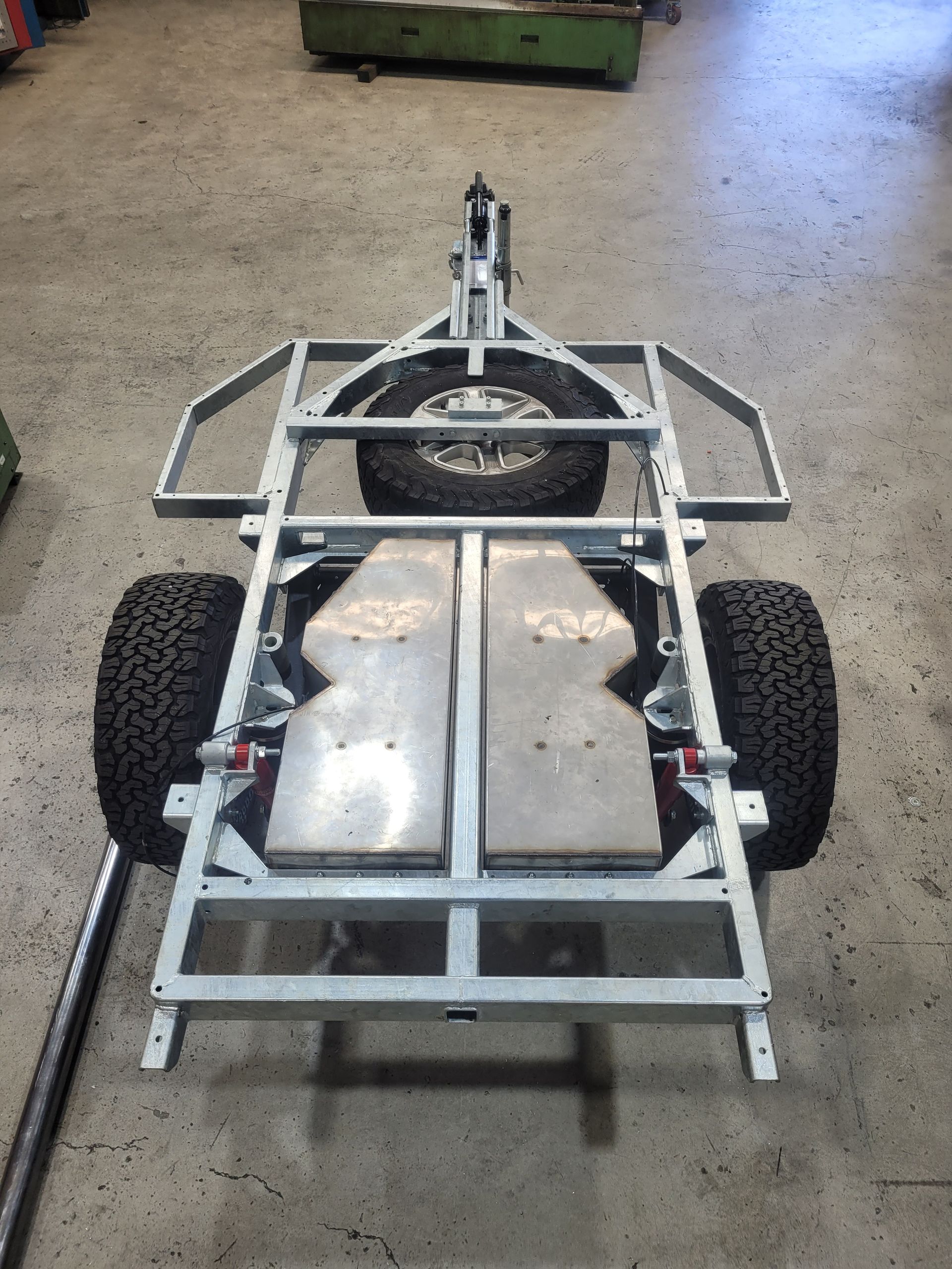 A galvanized steel trailer frame with two all-terrain tires, a spare wheel, and metal floor plates in a workshop.