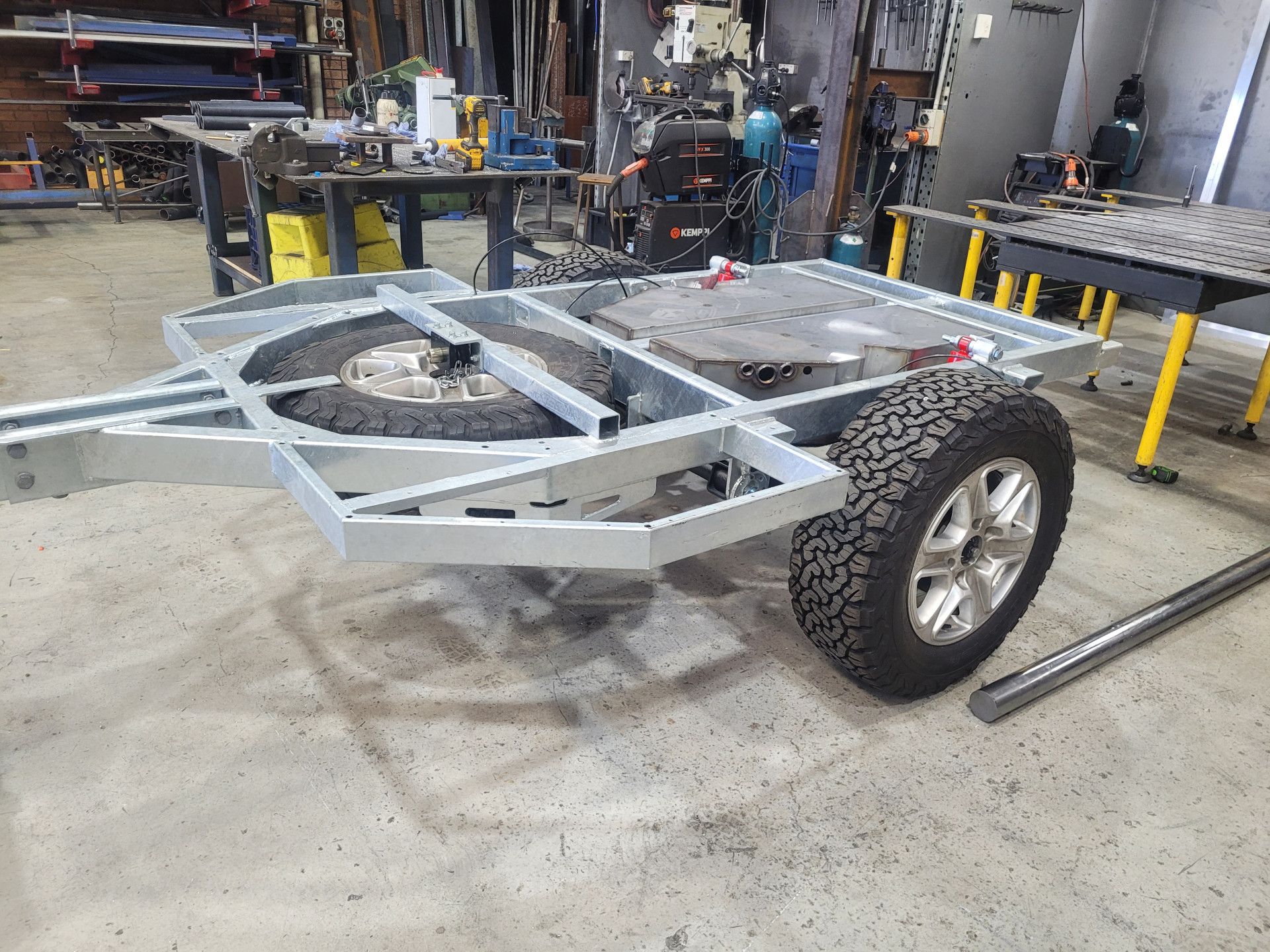 A galvanized metal trailer frame with a spare tire and wheel mounted on a concrete floor in a workshop.