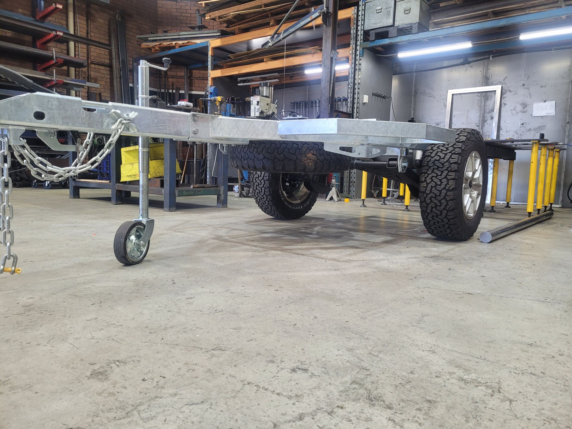 A galvanized metal trailer chassis with a jockey wheel and off-road tires in a workshop setting.