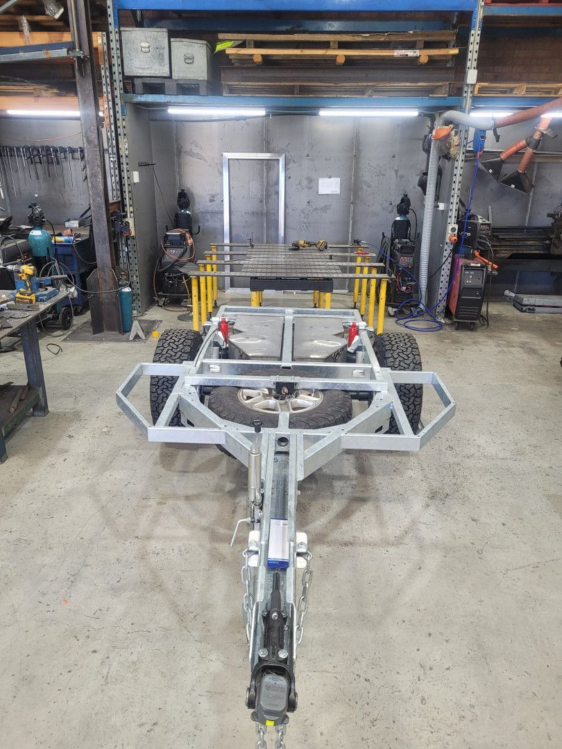 A metal trailer frame with two wheels and a spare tire sits in a workshop with concrete floors and industrial tools.