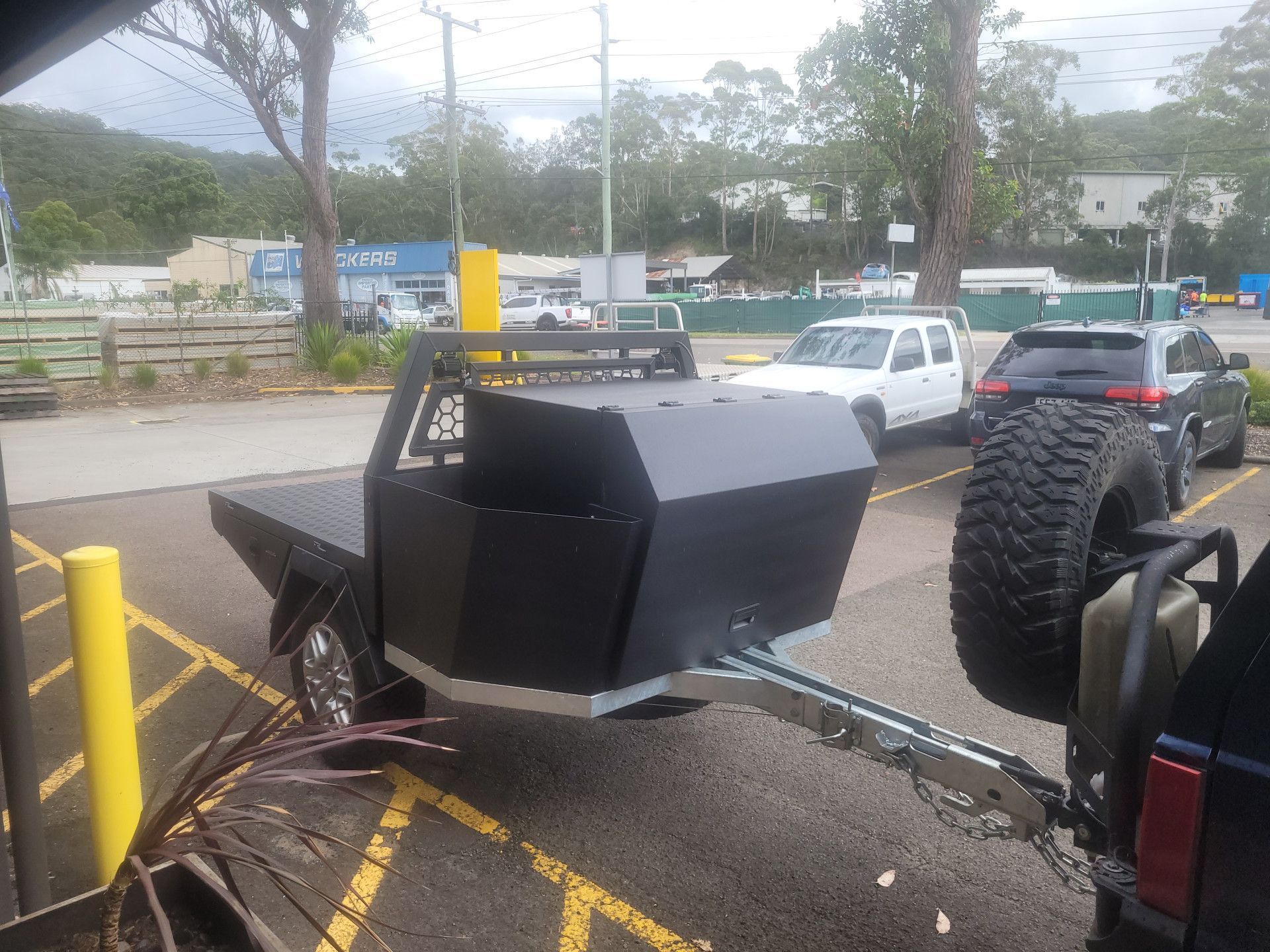 A black custom utility trailer with a storage box, parked in a lot behind a vehicle with a spare tire.
