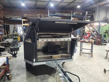 A metal storage box trailer with its top hatch open, sitting in a cluttered industrial workshop setting.