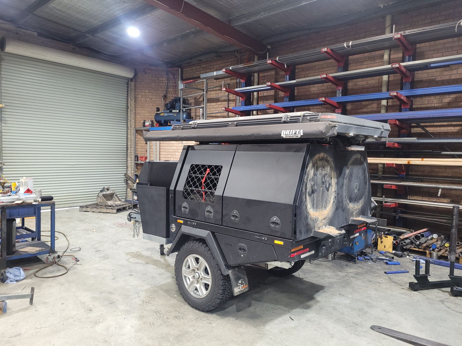 A black off-road camper trailer parked inside a workshop with storage racks and a roll-up garage door in the background.