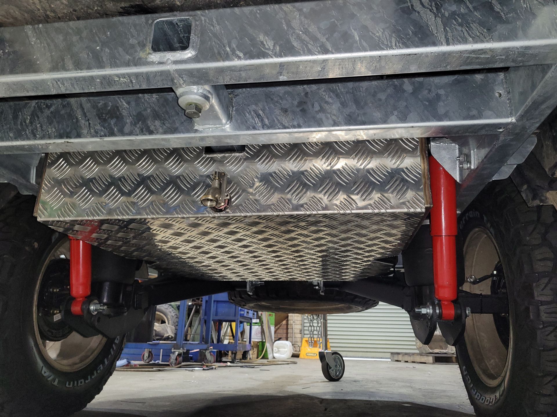 Underside of a vehicle showing an aluminum storage box mounted between two red shock absorbers and large tires.