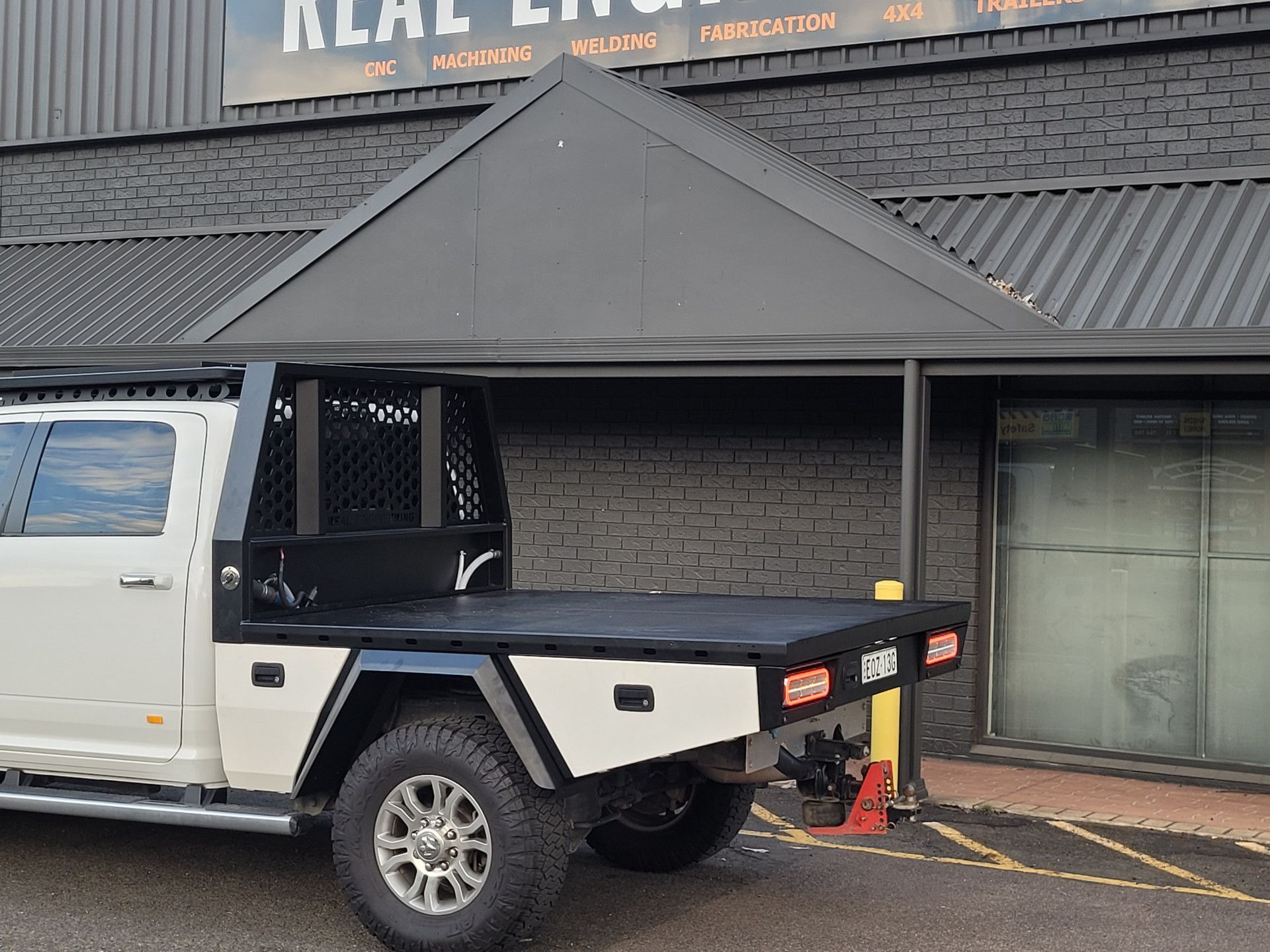 A white pickup truck with a custom black metal tray and headache rack parked outside an industrial building.