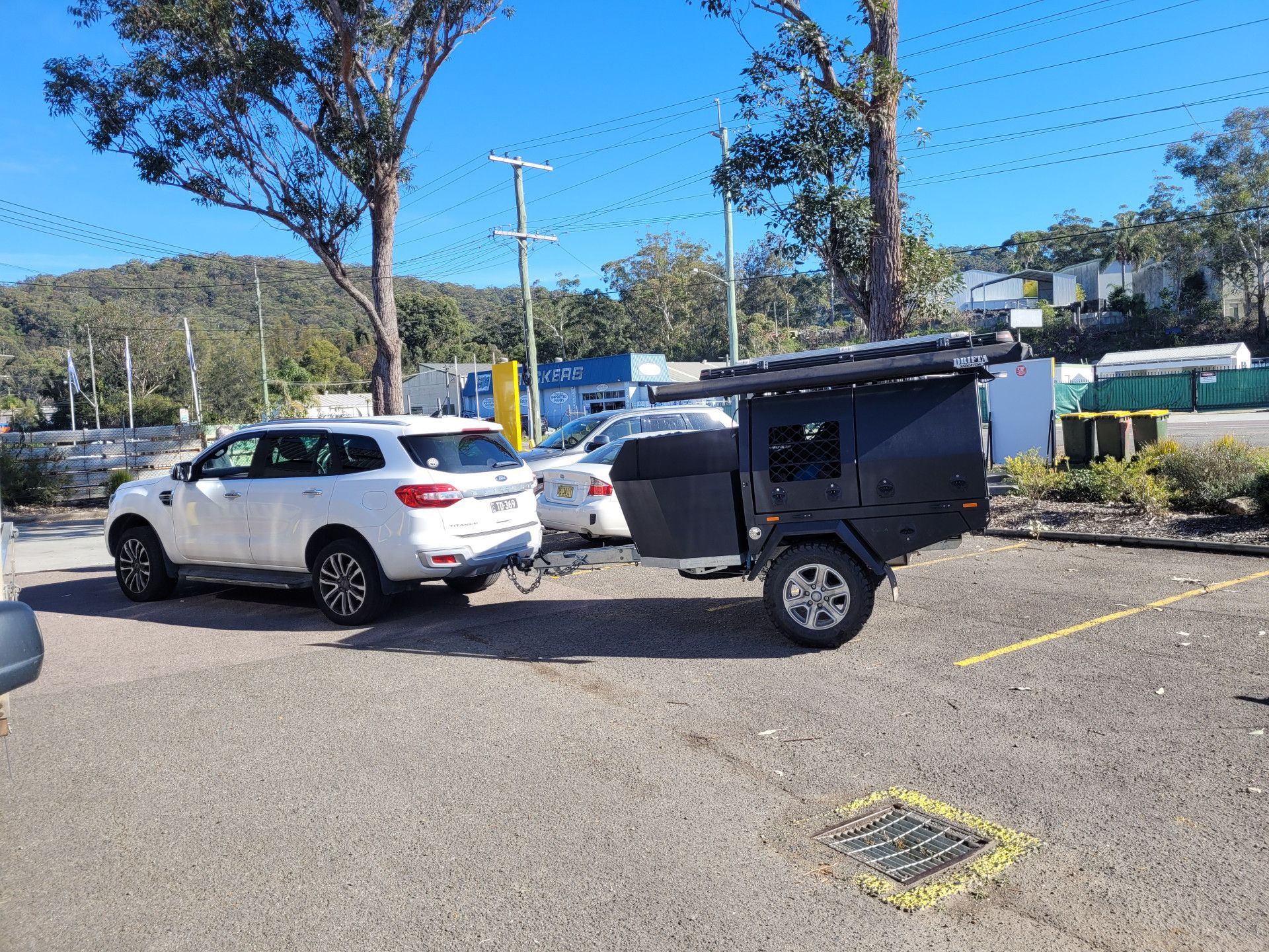 A white SUV parked in a gravel lot, towing a small, black box-style camping trailer.