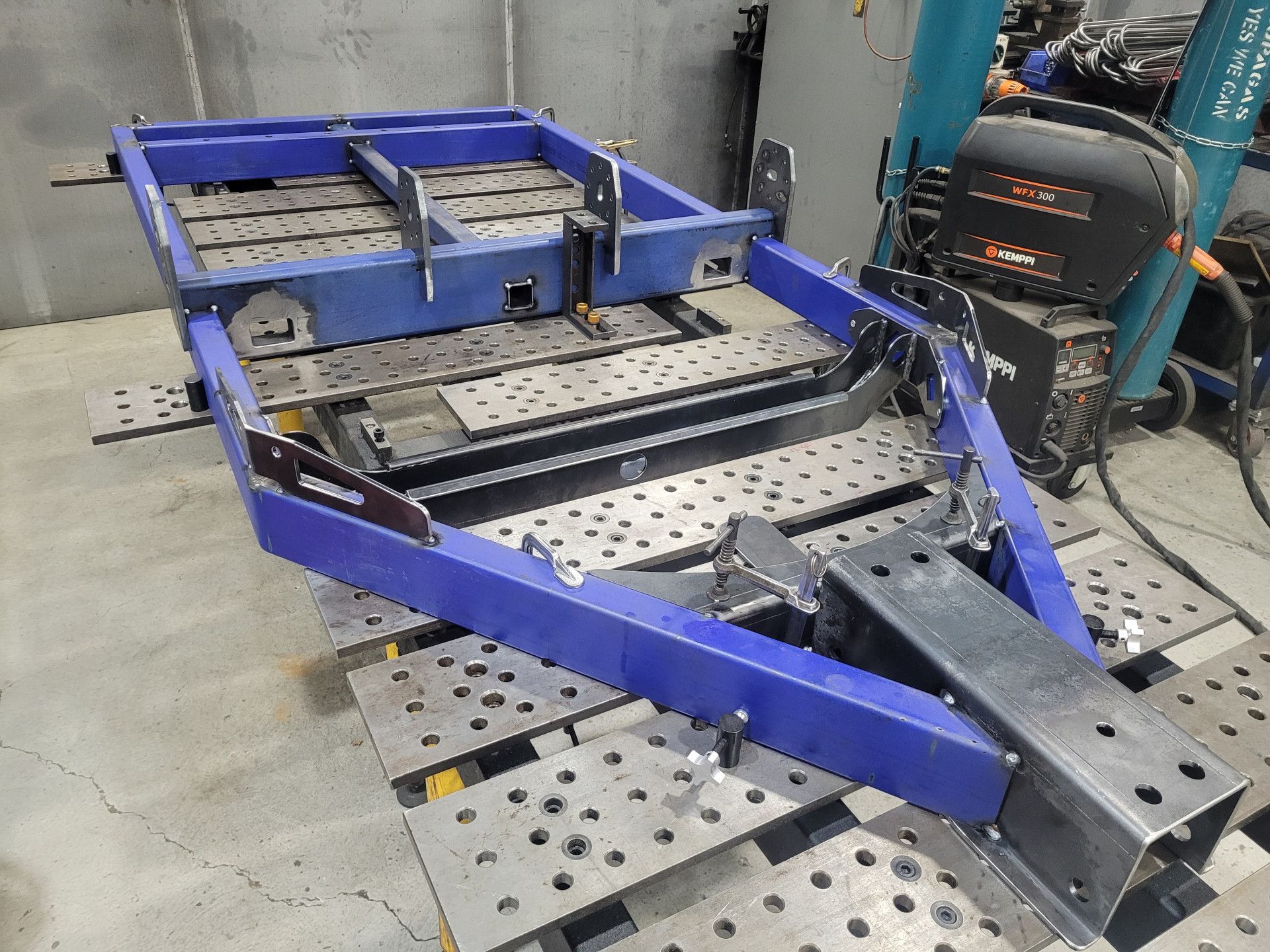A blue metal trailer frame under construction sits on a large, hole-patterned steel welding table in a shop.