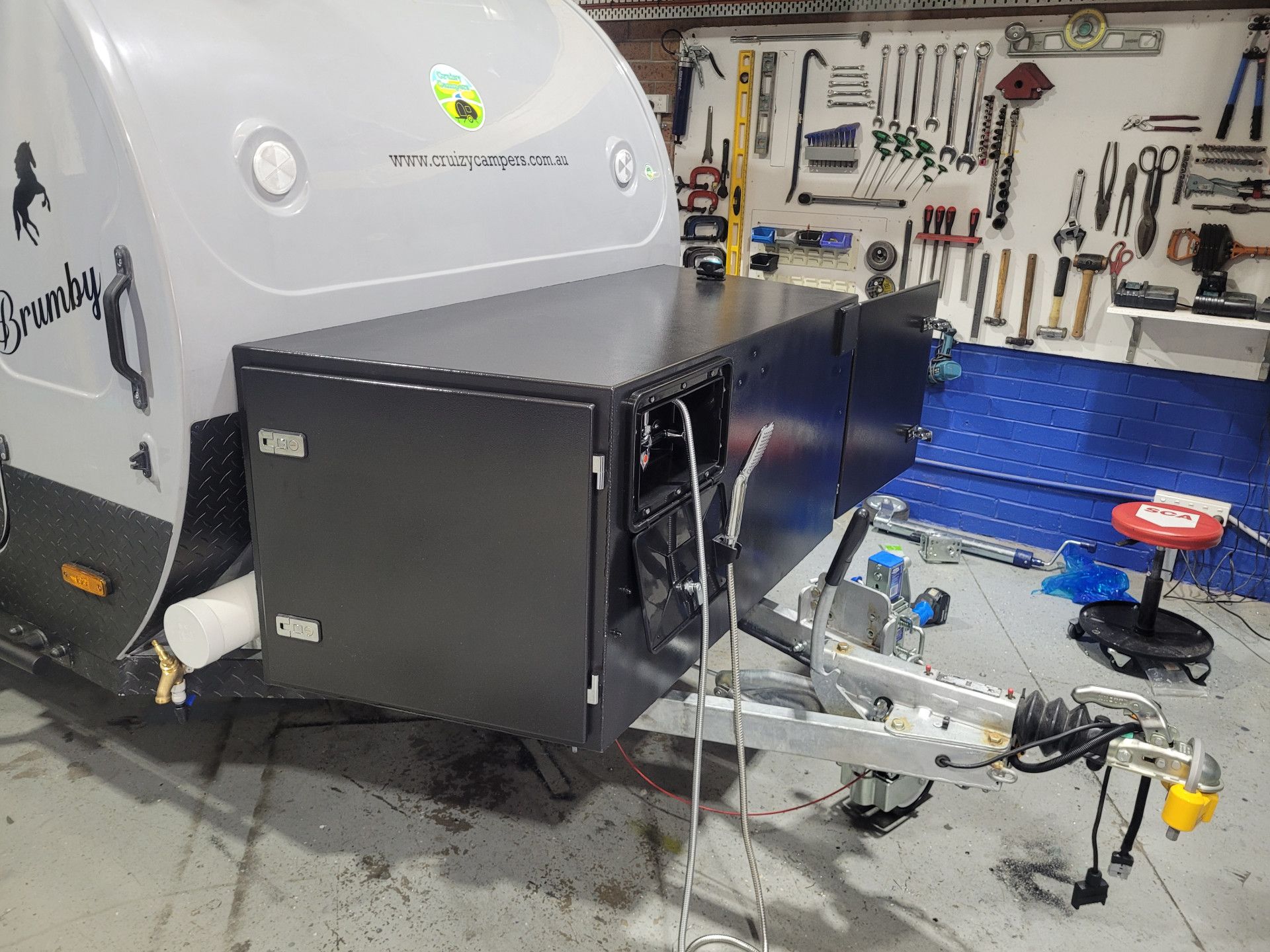 A close-up of a black storage box mounted on the front of a white camper trailer inside a workshop with tools on the wall.
